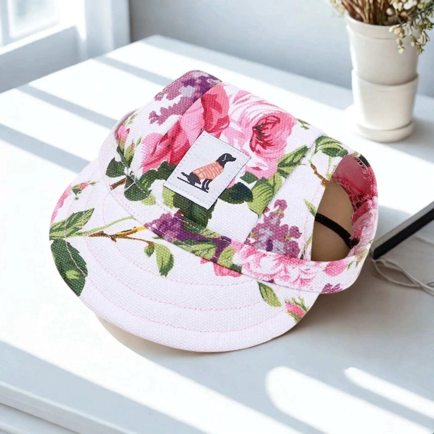 Une casquette chien fleuri rose sur un bureau blanc avec un petit pot de fleurs.