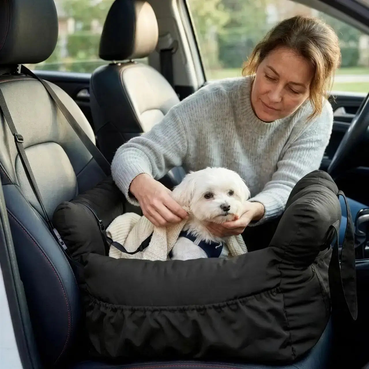 Une femme avec son bichon frisé dans son siege d'auto pour chiens noir.